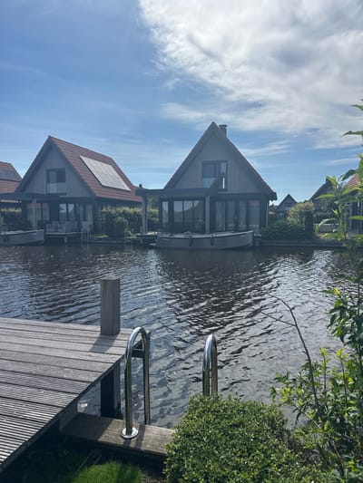 The image depicts a waterside scene in Ossenzijl, Netherlands, under a partly cloudy sky. In the foreground, a wooden pier extends into dark, rippling water. A metal ladder with two handrails is attached to the pier, descending into the water. Lush green bushes and foliage are visible to the right of the pier.
Across the water, several modern houses with red tile roofs are situated. These houses are characterized by large windows and wooden facades. Some of the roofs are equipped with solar panels. Two small boats are docked in front of the houses. The overall environment suggests a tranquil residential area adjacent to a canal or body of water. There are no people visible in the image.