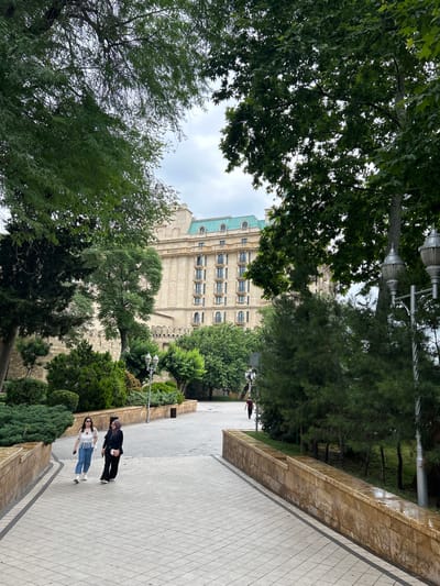 The image captures a sloped, paved walkway with a stone wall bordering it on the left. Two women are walking up the walkway. The woman in front is wearing a white shirt and blue jeans, and the woman behind her is wearing a black jacket and pants. Both are casually dressed and appear to be strolling. The walkway is lined with manicured bushes and trees, creating a park-like atmosphere. In the background, a large, ornate building with multiple windows and a greenish roof is visible, partially obscured by trees. The sky is overcast, suggesting a cloudy day, possibly late afternoon or early evening given the diffused light. The overall scene depicts a peaceful moment in a public outdoor space, likely a park or a promenade in an urban setting. The location context places this scene in Sabail Raion, Azerbaijan.