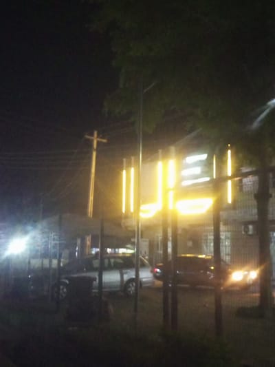 This is a nighttime photograph taken in Baccure Kofare, Nigeria. The scene appears to be a street or an area with a building and parked vehicles. The lighting suggests it's after dark, with artificial lights illuminating the area.

The main subjects are several vehicles. In the foreground, a dark-colored sedan with its headlights on is visible, parked behind some vertical poles. To its left, a lighter-colored sedan or SUV is parked. Further to the left, there's a blurred bright light that could be from another vehicle or a street lamp. The background shows a building with bright, yellow vertical lights, possibly signage, and some windows. A utility pole with wires is also visible in the middle ground. Trees with dark foliage are present, partially obscuring the sky and the top of the building.

There are no people clearly visible in the image. The overall mood is somewhat dark and moody due to the nighttime setting and the limited visibility. The presence of vehicles suggests it could be a public area, perhaps near shops or residences. There is no visible text that can be deciphered. The weather is not discernible from the image.