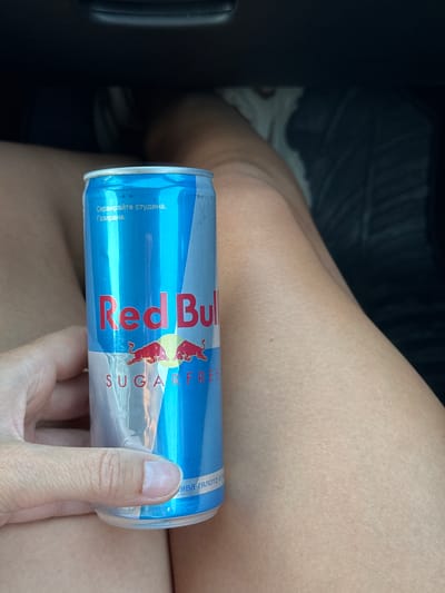 A hand holds a can of Red Bull Sugarfree. The can is blue with the Red Bull logo and text. The background shows a person's legs and a dark interior, possibly a car. There is no information available about the city of Churek, Bulgaria, or any specific location.