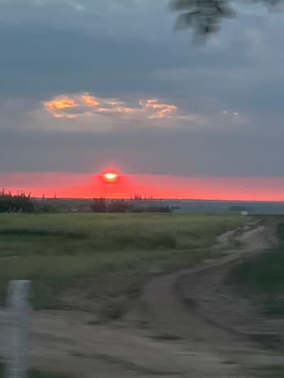 The image shows a sunset scene, likely over the countryside or a relatively open area. On the left is the sky with clouds that appear to be reflecting the setting sun's light. In the middle of the image, the sun is visible as a bright orange-red disk, partially obscured by the horizon. The horizon is lined with some indistinct elements that may be trees or distant buildings. To the right of the horizon, there is land, possibly a field or undeveloped terrain, with various shades of green and brown. The image is oriented sideways, with the sky and clouds on the left and the land on the right. There is no evidence to confirm the location is Karagash, Moldova.
