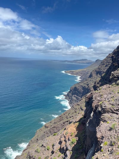 The photograph captures a coastal scene in La Aldea de San Nicolás, Spain, featuring rugged cliffs plunging into the Atlantic Ocean. The cliffs, composed of dark volcanic rock with sparse vegetation, dominate the right side of the image. Waves crash against the base of the cliffs, creating white foam. The ocean extends towards the horizon, its surface varying in shades of blue. A coastline of distant cliffs stretches into the background. The sky, visible on the left, is a mix of blue and white, with scattered clouds. The photo is taken from an elevated perspective, likely from a vantage point on the cliffs.
