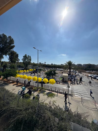 The image captures an overhead view of a large outdoor gathering in Kfar Yona, Israel, under a bright, clear blue sky with intense sunlight visible in the upper right quadrant. A substantial crowd, predominantly dressed in white, is densely assembled in an open, unpaved area. Along the metal crowd control barriers separating this gathering from a paved sidewalk and road, multiple clusters of bright yellow balloons are prominently displayed. Numerous individuals within the main crowd are also observed holding yellow objects or balloons.
On the paved areas, several individuals in darker clothing are seen walking or standing, including one person in what appears to be a dark uniform near a marked crosswalk in the lower right. Two scooters are parked on the sidewalk in the foreground, one equipped with a teal delivery box. Various vehicles are parked in surrounding areas, including multiple cars in lots adjacent to the gathering. The environment features several trees and shrubs, notably a prominent palm tree situated within the crowd area, and dense foliage along the left side. A small, light-colored rectangular structure is also present amidst the gathering. Distant residential buildings are visible on the horizon to the upper right. Streetlights are present throughout the scene.