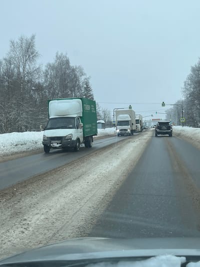 This image depicts a winter road scene in Borisovo, Russia, under an overcast, light gray sky. The asphalt road shows two visible lanes in the direction of travel, with a thin layer of dirty snow and slush accumulated on the shoulders and outer edges, while the main travel paths are clearer. Snow-covered, bare deciduous trees and some evergreens line both sides of the road.
In the left foreground, a white box truck, possibly a GAZelle model, with a green cargo box bearing the Cyrillic text "ГЕЙЗЕР" and other smaller text, is visible. Its license plate, "H632AE 50", is partially obscured but legible. This vehicle appears to be moving or stationary in the same direction as the camera.
In the mid-ground, a line of vehicles proceeds or waits. The most prominent is another white box truck with its headlights illuminated, followed by at least two other white box trucks or cargo vans. Further down the road, a dark-colored SUV is visible.
Overhead, multiple traffic lights are visible in the distance, displaying green signals. Yellow, diamond-shaped pedestrian crossing signs are mounted on poles above the road. On the right shoulder, a structure resembling a bus stop shelter, partially covered in snow, is present. The bottom edge of the image shows the dashboard and a small amount of snow on the windshield of the vehicle from which the photograph was taken. No identifiable people are visible.