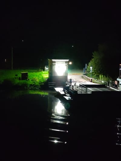 The image shows a nighttime scene of a cable ferry reflected in water, likely a river or canal. On the far bank, there's a small building emitting bright light, which is also reflected in the water. The surrounding area includes a grassy embankment and some trees on either side. The scene suggests it might be a rural or semi-rural location, and the cable ferry is the central element. The scene shows the Lesnoi cable ferry at night.