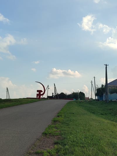 The image depicts a rural scene in the village of Karabashyovo, Russia, under a bright blue sky with scattered white clouds. A paved road curves into the distance from the foreground, bordered by a grassy embankment on the right. On the left side of the road, on a slightly elevated mound, stands a prominent red sculpture resembling a hammer and sickle. Several utility poles with power lines are visible along the road and in the background, some marking the edge of a residential area. In the distance, to the right of the sculpture, are houses with blue roofs and fences, and a stand of trees. A small flock of sheep is grazing on the grassy area in front of the houses. The overall atmosphere is serene and typical of a village setting.