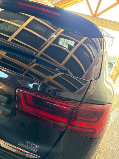The image depicts the rear exterior of a dark-colored vehicle, likely an Audi, parked outdoors in Paisievo, Bulgaria. The primary focus is the left-rear quarter of the vehicle, showcasing its polished surface, illuminated red LED taillights, and "TDI quattro" badging. A rear wiper blade is visible on the dark rear window. The highly reflective surface of the vehicle, particularly the rear window and upper bodywork, displays a clear reflection of an overhead structure composed of light-colored wooden beams forming a grid pattern. Reflections of light-toned building materials, possibly brick or concrete walls, are also present on the right side of the vehicle's bodywork. No individuals, actions, or interactions are discernible within the frame. The scene is brightly lit, suggesting daylight conditions.