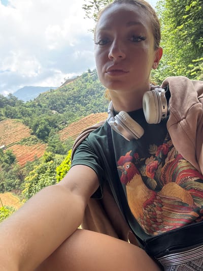 A woman is seen from an upward angle, likely a selfie, in Hoàng Su Phì Commune, Vietnam. She wears headphones around her neck and a dark green shirt with a large rooster design on the front. The background features lush green mountains and terraced fields. The sky is partly cloudy.