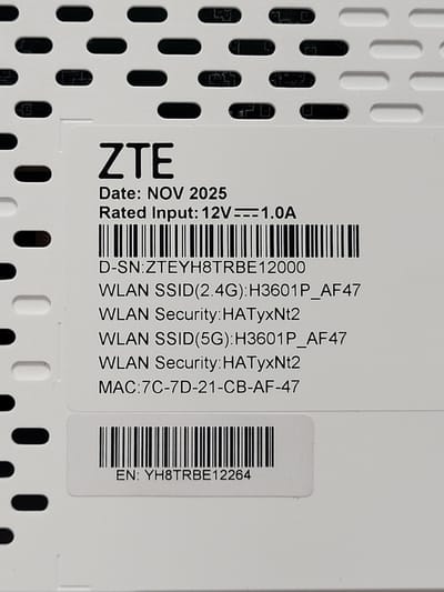 The image displays a close-up, top-down view of a white rectangular product label affixed to a white, perforated electronic device. The label features the brand "ZTE" in prominent black text at the top, followed by "Date: NOV 2025" and "Rated Input: 12V === 1.0A". Below this, a black linear barcode is present, identifying the serial number "D-SN:ZTEYH8TRBE12000". Further details include "WLAN SSID(2.4G):H3601P_AF47", "WLAN Security:HATyxNt2", "WLAN SSID(5G):H3601P_AF47", "WLAN Security:HATyxNt2", and "MAC:7C-7D-21-CB-AF-47". A second, shorter black linear barcode is visible near the bottom of the label, corresponding to "EN: YH8TRBE12264". The background of the label itself is white. The underlying electronic device is white with a pattern of horizontally elongated, oval-shaped perforations, consistent with a ventilation grille. These perforations are dark internally, suggesting depth. No people, discernible actions, or interactions are visible. The scene is an inanimate object in a close-up setting. The specific city location in the United States of America is not discernible from the image.