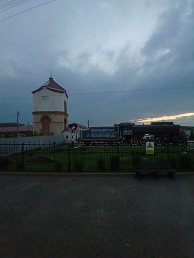 An outdoor, low-angle, medium shot captures a historical monument in Abdulino, Russia. The scene depicts a preserved steam locomotive, identified as "Л-77", positioned alongside a water tower. The locomotive, painted dark, stands on a section of railway track and is accompanied by a plaque inscribed with Russian text. The water tower, a brick and concrete structure with a conical roof, is situated to the left of the train. A black metal fence encloses the monument area, which is landscaped with green grass. In the foreground, a dark asphalt surface and a wooden bench are visible. The sky above is overcast with dramatic clouds, suggesting twilight or a stormy weather condition. No individuals are present in the frame. The image is taken from a street or pathway looking towards the monument.
