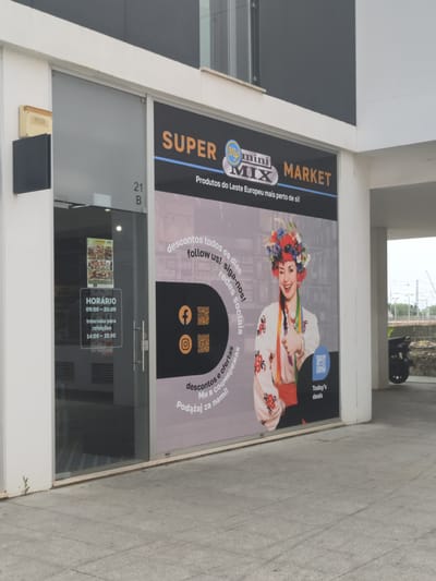 An outdoor, eye-level shot captures the modern storefront of "SUPER mini MIX MARKET" in Algés, Portugal, during daylight hours. The market specializes in "Produtos do Leste Europeu mais perto de si!" (Eastern European Products closer to you!).

The main display window features a vibrant graphic of a smiling, winking woman wearing a colorful, traditional Eastern European floral headdress and embroidered blouse, giving a thumbs-up. To her left, text in Portuguese and other languages (likely Ukrainian and Polish, based on the market's focus) encourages customers to "follow us!" and highlights "descontos todos os dias" (discounts every day) and "descontos e ofertas" (discounts and offers). Social media icons for Facebook and Instagram are displayed with associated QR codes. Another QR code near the woman is labeled "Today's deals".

The adjacent glass entrance door displays the operating hours: "HORÁRIO 09:00-20:00" with an "Intervalo para refeições 14:00-15:00" (meal break). A small poster, likely showcasing food items, is visible on the door. Above the door, the address "21 B" is marked. The building facade is a mix of white and dark gray panels. A small external fixture is mounted on the wall next to the entrance. The ground in front of the store is paved with light gray tiles, with a small green sprout emerging from a crack near the base of the wall. The scene suggests a welcoming commercial establishment.