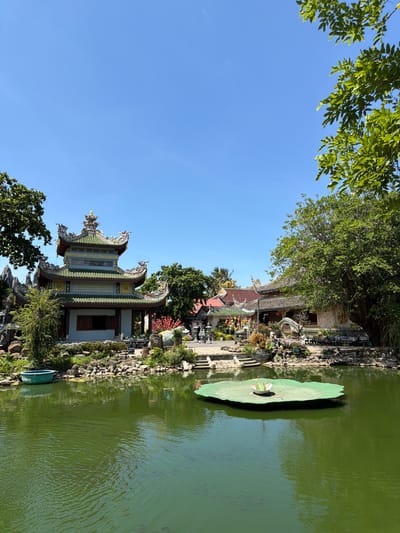 The image shows a scenic view of a temple complex with a pond in the foreground. The temple buildings feature traditional East Asian architecture with ornate roofs and intricate decorations. The pond is filled with murky green water and contains several fish. In the center of the pond is a large, artificial lily pad with a frog-shaped decoration. The surroundings include lush trees, rocks, and flowering plants. The sky is clear and blue. The location is identified as the Thien An Temple in the city of Hòa Đa, Vietnam. There are no people visible in the image.