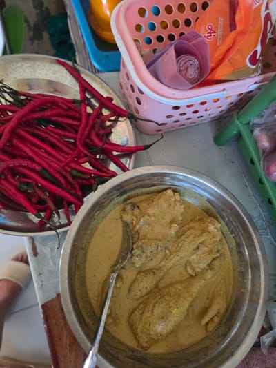 The image depicts a still life of food items and household objects on a table, potentially in Pintomakmur, Indonesia. In the foreground, a stainless steel bowl contains pieces of chicken immersed in a thick, yellowish curry sauce. A metal spoon rests inside the bowl, partially submerged in the sauce. To the left of the bowl, a silver platter is filled with a pile of long, vibrant red chilies, some with green stems still attached.
In the background, a pink plastic basket holds various items, including a rolled-up pink plastic sheet and an orange packet with text. To the right of the pink basket, a green plastic container is visible. The table surface appears to be a light-colored laminate or formica, with visible wear and tear. A partial view of a person's leg wearing a light-colored slipper is in the bottom left corner of the image, suggesting a domestic setting. The overall scene conveys a sense of everyday life and food preparation.