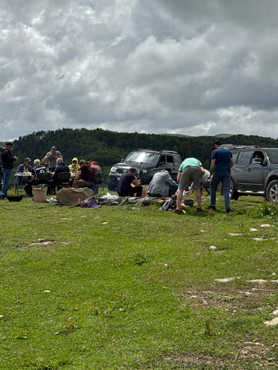 In Nizhnyaya Teberda, Russia, a group of people are gathered in a grassy, outdoor area, likely for a picnic or a recreational outing. The scene is dominated by lush green grass and a backdrop of rolling hills covered in dense, dark green trees. The sky above is overcast with dramatic, grey clouds. Two large, dark-colored SUVs are parked prominently in the middle ground, suggesting transportation to this remote location.

In the foreground, several individuals are engaged in various activities. A table is set up with people seated around it, some appearing to be eating or conversing. Nearby, a campfire is visible with logs and what appears to be purple fabric scattered around it, indicating recent or ongoing activity. Other individuals are standing or bending over, some near the campfire and others near the vehicles, possibly preparing food or organizing gear. The overall impression is of a casual gathering in a natural setting.
