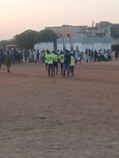 A sports team wearing light green jerseys with dark numbers stands on a brown field. Spectators stand along the edge of the field and in the background. Behind the spectators are trees and low buildings. The sky is light and hazy.