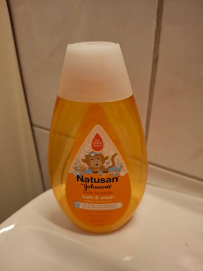 A close-up shot shows a clear, orange-tinted bottle of Natusan Kids Bubble Bath & Wash, a Johnson's product, sitting on a white surface, likely a sink or countertop. The bottle has a white cap and a label with a cartoon monkey and bubbles. A red droplet symbol with the text "no more tears" is also visible on the label. The background is a textured, light brown wall. There are no people or animals present. The scene is indoors, in what appears to be a bathroom. There is no information to place this image specifically in Åsgårdstrand, Norway.