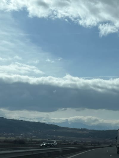 The image depicts a highway scene within a mountainous landscape, identified as the vicinity of Vladimir, Bulgaria. The sky is partially cloudy, featuring patches of clear blue above a prominent horizontal band of dense, dark grey clouds, which are topped by brighter white cloud formations. Two faint aircraft contrails are visible in the upper and mid-sky. In the background, a distant mountain range with snow-capped peaks extends beneath the cloud cover. Closer hillsides are covered in dark green and brown vegetation, with a small settlement visible on the lower slopes to the left.

In the midground, a multi-lane asphalt highway with a grey metal median barrier is present. On the left side, two dark vehicles are observed traveling in the opposing direction: a dark SUV, possibly a Jeep, followed by a smaller dark car. On the right side, the front portion of a white semi-trailer truck is visible, moving in the same direction as the camera, with another white vehicle further ahead in the right lane. The road surface is dark asphalt and appears dry. No individuals are discernible.