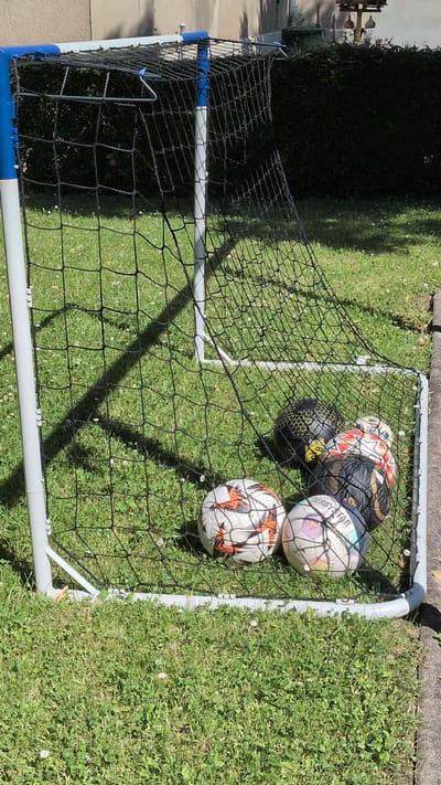 The image shows a small, portable soccer goal net in an outdoor setting, likely a backyard or park, given the presence of green grass and trees in the background. The goal net is made of white PVC pipes and black netting. Inside the net, there are five soccer balls. One ball, prominently in the front right, is orange and white with a distinct panel pattern. To its left and slightly behind is a white ball with colorful geometric patterns, and the text "DERBYSTAR" is visible on it. Behind these, there are three more balls: a black and yellow ball with a hexagonal pattern and a "BVB" logo, a black and gold ball, and a red and white ball with a similar panel pattern to the orange and white one.

The sun is casting distinct shadows on the grass, suggesting it is daytime. The lighting indicates a bright, sunny day. The overall scene is one of leisure or practice, with the soccer balls ready for play. There are no people visible in the image. The setting appears to be a well-maintained grassy area, possibly in Buttstädt, Germany, as indicated. The image is taken from a slightly elevated, overhead perspective, looking down into the soccer goal.