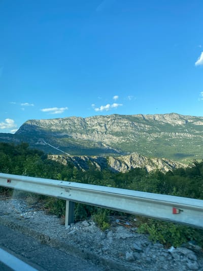 The image shows a scenic view of a mountain range with a bright blue sky. In the foreground, a metal guardrail runs horizontally across the frame, separating the road from a steep drop-off covered in lush green vegetation. The road itself is visible at the bottom of the image, with gravel and debris on its surface. The mountain range dominates the background, with rugged, rocky slopes interspersed with dense green foliage. A long, white pipe or cable is visible snaking its way up the mountainside, suggesting some kind of infrastructure or water management system. The overall impression is one of a natural landscape, likely in a mountainous region, with evidence of human presence in the form of the guardrail and the pipe. While the exact location is not specified, the scene evokes a sense of being on a scenic drive or hike in a mountainous area.