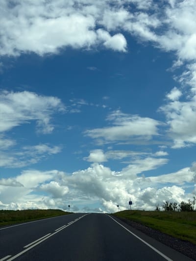 The image shows a highway in the city of Ostrovnoye, Russia, stretching towards a hill under a bright blue sky filled with fluffy white clouds. The road is paved and has double white lines marking its lanes. To the left of the road, there is a grassy field. To the right, there is a grassy embankment with some trees and bushes at the top of the hill. Two road signs are visible on the right side of the road. One is a circular sign with a white border on a dark background, and the other is a blue rectangular sign with the number "70" in white, indicating a speed limit. The scene appears to be taken from a vehicle moving along the road. There are no people or animals visible in the image.