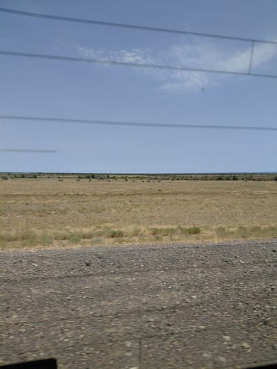 The image shows a wide, flat, open landscape under a clear blue sky with some scattered clouds. The ground is covered with dry, golden-brown grass and scattered bushes. In the distance, a line of low, dark green trees and vegetation marks the horizon. The foreground shows a gravelly surface, possibly a road or path. Horizontal lines, suggestive of window blinds or bars, are visible across the upper portion of the image, indicating the photo was taken from inside a vehicle or building. The scene is devoid of any people or animals. The location is identified as Ынталы, Kazakhstan.