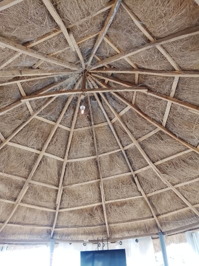 This image presents an upward view into the interior of a conical thatched roof structure, located in Farin Yashe, Nigeria. The roof features a visible internal framework of radial wooden rafters converging at a central point and interconnected by horizontal wooden battens, forming a grid. Dried straw or thatch material fills the sections between the wooden supports. An illuminated incandescent light bulb hangs by a wire from the apex of the roof. Below the lower edge of the thatch, the scene reveals a bright, possibly white, wall or curtain system. A dark rectangular screen, consistent with a television, is mounted below the roof structure in the lower central portion of the frame. Several metallic rings or hooks are visible along the bottom perimeter of the roofline. A partial "NO SMOKING" sign is faintly visible on the lower left. No individuals are present in the scene.