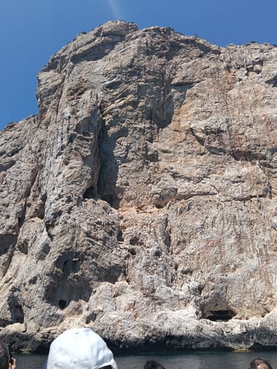 This is a wide-angle, ground-level photograph taken from a boat on the water, looking up at a large, rugged, light-colored rock face. The rock is textured with many crevices, ledges, and some darker shadowed areas, including a prominent cave entrance towards the center-right of the frame. The rock face appears to be a cliff or a significant geological formation. The sky above is a clear, bright blue. In the foreground, at the bottom of the image, the heads and shoulders of at least three people are visible, suggesting they are passengers on the boat. One person in the center is wearing a white baseball cap. The water is dark and appears relatively calm. This cliff is likely located along the coast near Alghero, Sardinia, Italy, possibly within or near the Capo Caccia - Isola Piana Marine Protected Area, known for its dramatic limestone cliffs and sea caves.