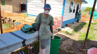 This media file captures an outdoor scene at the Hôpital Général de Référence Rwampa in Tchere, Democratic Republic of the Congo. The setting features simple buildings, unpaved ground, and orange safety fencing, suggesting a temporary or field medical facility.

In the foreground, a healthcare worker stands behind a table covered with a white tarp. They are fully clad in personal protective equipment (PPE), including a blue cap, a white face mask, a clear plastic face shield labeled "Face Shield Direct Splash Protection," green scrubs, a clear plastic apron, and white gloves. On the table, several neatly folded green garments, likely additional scrubs or protective gowns, are stacked. A green plastic waste bin and a stainless steel thermos are visible near the table.

In the mid-ground, another individual in green scrubs stands near a white and blue painted wooden building. Clothes are seen hanging outside this building. Behind the orange fencing, various pieces of footwear, including boots and sandals, are placed on wooden posts, possibly for drying or storage. To the left, another building with textured, possibly mud, walls is visible.

The scene unfolds during bright daylight under clear skies. The visible text on the white building, "HOPITAL GENERALE DE REFERENCE RWAMPA" and "FSFACE SUR," confirms the location as a medical facility. The overall impression is of a functional, albeit basic, healthcare operation, emphasizing readiness and infection control in a resource-limited environment. A watermark "Joseph Kisuki" is present in the lower right corner.