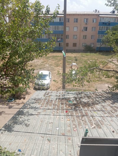 A street-level exterior shot captures a multi-story apartment building in Nurabad, Uzbekistan, on a bright, sunny day. The building has a light brown facade with blue accents around windows and balconies. Several windows are open, and laundry is visible hanging from a balcony on the right side. In front of the building, a patch of dry, mowed grass is visible. A white car is parked on a paved area to the left of the building, partially obscured by a leafy green tree. A utility pole stands in the foreground, with a "Wi-Fi" sticker attached. Beyond the grassy area, a second, lower structure with a corrugated metal roof, covered in leaves and debris, is in the foreground. The roof is also adorned with several pieces of trash, including plastic bags and wrappers, as well as two green clothespins. The scene suggests a residential area, possibly in a slightly neglected state, given the debris on the lower roof.