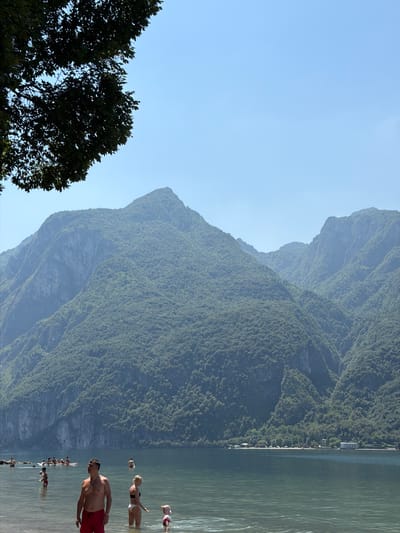 The image depicts a sunny day at a lakeside with a stunning mountainous backdrop. The scene is set in Abbadia Lariana, Italy, as suggested by the landscape. The mountains are covered in lush greenery, with a clear blue sky above. The lake itself is calm and reflects the surrounding scenery.
Several people are enjoying the water. In the foreground, a man with dark hair and a tan torso is wading in the lake, wearing red swimming shorts. To his right and slightly further back, a woman with blonde hair is wearing a bikini. Further in the distance, other individuals are visible in the water, some appearing to be swimming or wading. The overall atmosphere suggests a recreational day spent by the lake, likely during warm weather given the attire of the people. The time of day appears to be daytime, with bright sunlight casting a natural illumination on the scene. There is no visible text in the image. The composition, with the towering mountains and the serene lake, creates a picturesque and inviting setting.