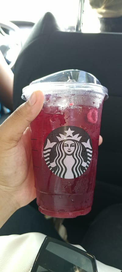 A person's hand holds a clear plastic cup filled with a dark red, cold beverage. The cup has condensation on the outside, indicating it is chilled. A black and white Starbucks logo, featuring the iconic siren, is visible on the cup. The beverage appears to contain fruit pieces. The background is blurred but suggests an indoor setting, possibly a car, with dark upholstery and a glimpse of a white surface in the foreground. The lighting suggests it is daytime.