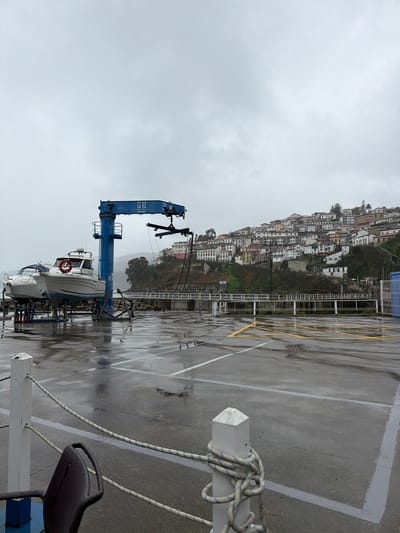 The image displays a port area in Colunga, Spain, under an overcast grey sky. The immediate foreground shows wet, dark asphalt pavement with visible reflections, marked by white and yellow painted lines indicating parking or designated zones. A white wooden bollard, wrapped with a blue and white striped rope, is present in the lower-left quadrant. A dark, possibly metal, chair back is partially visible in the bottom-left corner.

In the midground, a prominent blue gantry crane or boat lift, marked "GE" and "GRUA 800", stands on the wet pavement. To its left, two white recreational boats are dry-docked on support stands. The boat closer to the crane features a red lifebuoy on its side and displays the registration number "7E-AM-1-18-01". The crane's lifting mechanism, complete with black slings, hangs suspended. Behind these structures, a white-railed bridge or walkway extends horizontally, with a visible stone or concrete retaining wall beneath.

The background features a steep, green hillside densely populated with numerous multi-story houses. These residences are predominantly white with varied roof colors, including red, brown, and orange, ascending the slope. Trees and vegetation are interspersed among the buildings and cover parts of the hillside. No individuals or active movements are observed in the scene.