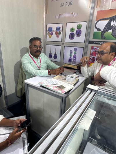 This image captures an interior scene within an exhibition booth at an event in Jaipur, India, as indicated by the prominent "JAIPUR" signage on a background display board.
Three adult males are present. In the center, a man with a mustache, glasses, and a red tilak on his forehead, wearing a light green collared shirt and a pink "BRC" lanyard, is seated behind a white desk. He has red and yellow beaded bracelets on his right wrist. Opposite him, on the right side of the desk, another man, bald and wearing glasses, a white kurta, and a pink lanyard, is seated. This man is actively gesturing with his raised right hand, engaged in conversation with the first man. In the bottom-left corner, only the hands of a third individual are visible, holding a black smartphone over a document with printed information.
The white desk separating the two main subjects holds various documents, including one labeled "Nan P", a small white compartmentalized box, and a partially visible scale. The background features display boards; one prominently shows four framed illustrations of intricate gemstone jewelry, including multi-colored, purple, and green earrings and pendants. An adjacent board displays a black tiger silhouette on an Indian flag background, with "MP" and "www.pbci.in" text. A glass display case with silver-colored rails is partially visible in the right foreground. The overall environment consists of modular light-colored partition walls and artificial lighting, characteristic of a trade fair or exposition booth.