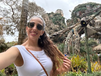 An adult female is depicted taking a selfie outdoors in Pandora – The World of Avatar at Disney's Animal Kingdom, located in Bay Lake, Florida, United States. The subject, in the foreground, has long, wavy, reddish-brown hair, wears hexagonal-framed sunglasses, a white sleeveless top, and a gold-colored necklace with an infinity symbol pendant. She is smiling at the camera, with her right hand, displaying pink and white polka-dotted fingernails, touching her hair. The background consists of intricate, themed environmental structures characteristic of Pandora, featuring large, gnarled trees, exposed root systems, and simulated "floating mountains" covered in lush green foliage. Several dark, flute-like architectural elements with straw caps and glowing yellow interiors are integrated into the scene. A tall, bare tree trunk stands on the left. The ground is covered with various green plants. A partially visible figure in blue is in the distant background. The sky is overcast.