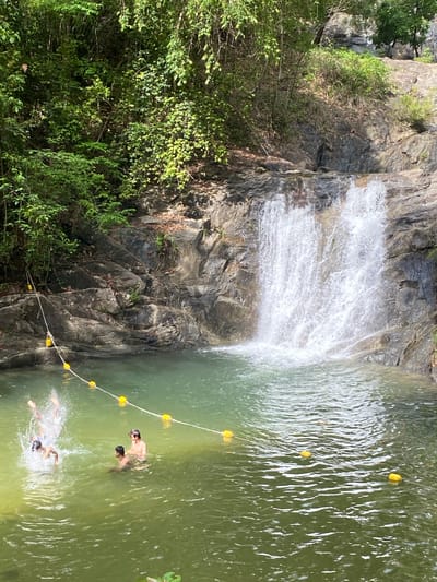 The image shows a waterfall cascading into a pool of green water, likely located in a natural area. The waterfall descends over a rocky cliff face partially covered with vegetation. A safety line with yellow buoys stretches across the pool. Three individuals are in the water: one swimmer is submerged and splashing, while two other individuals are partially submerged, standing or treading water near the buoy line. The surrounding environment consists of lush green foliage and rocky terrain, indicative of a natural, possibly mountainous location. The scene is captured in daylight. Based on the prompt, the location is likely in Tambon Thai Mueang, Thailand.