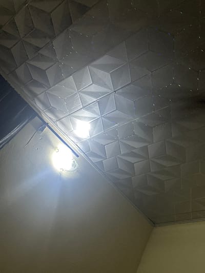 A textured, geometric-patterned ceiling occupies much of the frame, seemingly composed of raised, interconnected cubes. A bright light fixture, partially obscured by a support rod, casts a strong glare against an adjacent, plain white wall. The lower portion reveals a dark, undefined space, possibly another wall or fixture, adding depth to the composition. The image's perspective is from below, angled upwards toward the ceiling and light source.