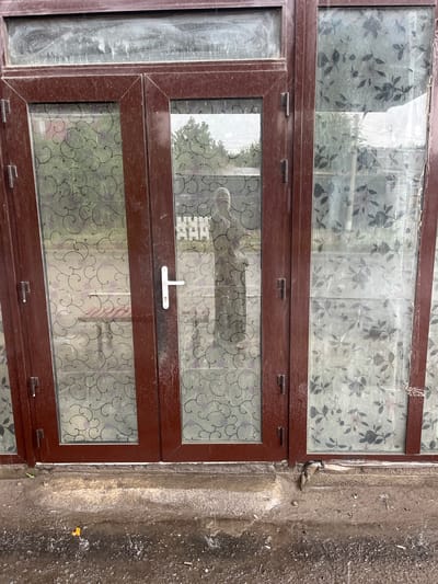 The image shows a close-up of a glass door with a brown frame, likely located in Medrese, Kyrgyzstan. The glass panels are decorated with a floral or swirly pattern, and there is a white door handle on the right-hand door. Reflections in the glass suggest an outdoor environment with trees and buildings. The bottom of the door frame appears to be resting on a rough, dirty surface, possibly ground or concrete, with some debris and water. The overall impression is one of an exterior entrance to a building, showing signs of weathering and some disrepair.