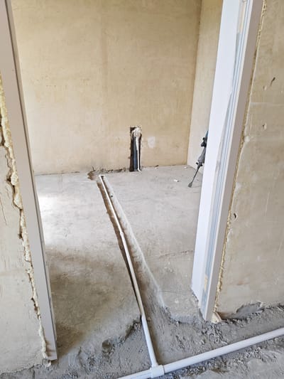 The image depicts an indoor construction scene in what appears to be a room under renovation. The subjects are plumbing pipes, a jackhammer, and unfinished walls and floors. The setting is a concrete-floored room with walls that are roughly plastered and show signs of construction work, including exposed concrete and sealant around door frames.

The primary activity shown is plumbing installation, with white PVC pipes laid in a trench in the concrete floor, running from a vertical pipe emerging from a wall cavity. A jackhammer, a tool used for breaking up hard surfaces like concrete, is visible leaning against a wall in the background, suggesting that the trench for the pipes was created or is being prepared.

Notable details include the raw, unfinished state of the room, indicating that the work is in progress. The lighting suggests it is daytime, and there are no indicators of weather conditions. The location cues are minimal, but the provided context states the location is Bazum, Armenia. There is no visible text in the image. The overall impression is one of active construction and renovation.