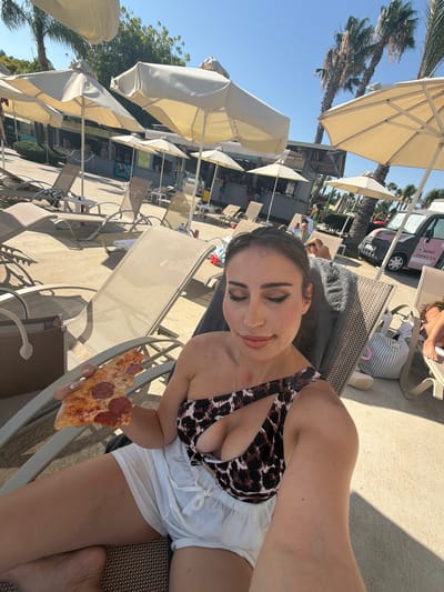 The image shows a woman sitting on a lounge chair by a pool, eating a slice of pepperoni pizza. She is wearing a leopard-print bikini top and white shorts. The sun is shining brightly, and the sky is blue, suggesting it is daytime and the weather is warm. Palm trees and white umbrellas are visible in the background, indicating a tropical or subtropical resort setting, consistent with the location context of Trachoni, Cyprus. Other lounge chairs and people can be seen in the distance, suggesting a public outdoor area like a resort pool. The woman has makeup on and appears relaxed, smiling slightly as she looks at the camera while holding the pizza. A small pink donut van is parked in the background, with text that reads "MINI DONUTS".