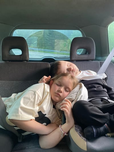 Two children are sleeping in the back seat of a car. The younger child, a boy, is on top, leaning his head on the older child's shoulder. The older child, a girl, is below, her head resting on a pillow. Both children are wearing casual clothing. The interior of the car appears to be dark grey fabric, and a silver seatbelt is visible. The background seen through the rear window is blurred, showing greenery and what appears to be a road. The scene is peaceful and suggests a long car journey. The image is taken from a slightly elevated angle, looking down at the children.