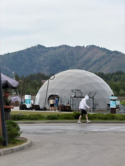 The image depicts an outdoor scene at a recreational facility with a large, geodesic dome structure. The dome, which appears to be a rental facility indicated by the Russian word "ПРОКАТ" (PROKAT) displayed on its side, is a prominent feature. In front of the dome, there are outdoor exercise equipment setups.
Several individuals are visible in the frame. A person wearing a white hooded jacket and black shorts is walking on a paved path in the foreground, moving from left to right. To the left of the dome, near a gray tent-like structure, a person in a green cap and green shirt is standing and looking towards the left, possibly a security guard or staff member. Two individuals are standing near the entrance of the dome, one wearing a light-colored backpack and the other in a blue t-shirt and shorts. Another person is partially visible sitting on a bench in front of the exercise equipment.
The environment includes a paved area, a grassy lawn with manicured bushes, and a backdrop of mountainous terrain covered with trees. The sky is overcast, suggesting a cloudy day. The overall scene suggests a public leisure or sports complex located in a natural setting, possibly in or near the city of Озерное (майминский), Russia, given the presence of Russian text.