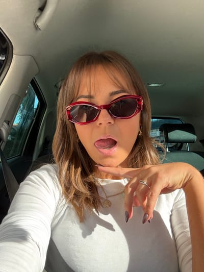 A female individual is depicted in a close-up selfie taken inside a vehicle in Vallauris, France, under bright sunlight. The subject has medium-length brown hair with bangs and wears red-framed sunglasses with dark lenses. She is dressed in a white, ribbed, long-sleeved top. Accessories include a gold-toned chain necklace with a circular pendant and a second, lighter-colored pendant, and a silver-toned ring on her left ring finger. Her fingernails are painted with a dark, gradient polish. The subject's mouth is open, with her tongue extended forward, and her left index finger rests against her chin. The vehicle's interior features a gray headliner, a visible gray grab handle on the left A-pillar, and a dark car seat headrest in the background to the right. Strong external light enters the vehicle, casting distinct shadows on the subject's top and the car's interior surfaces.