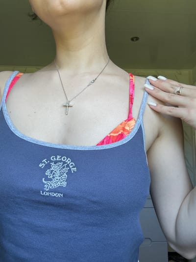 The media file shows a close-up, top-down view of a person's upper chest, shoulder, and arm. The person is wearing a dark blue tank top with a light grey trim and a bright pink and orange patterned bikini top underneath. A delicate silver necklace with a cross pendant is visible on the person's chest. On the person's left hand, which is resting near their shoulder, there are two rings on the ring finger and a manicure with white nail polish. The background is a muted green wall with a small, dark circular object that appears to be a light switch or vent. The lighting suggests it is daytime. The clothing features an embroidered crest on the tank top with the text "ST. GEORGE" and "LONDON." The overall impression is casual, possibly taken in a home or dressing room setting.