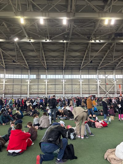 The image shows a large group of people seated on a green surface in a vast indoor space. The space appears to be an indoor arena or gymnasium, suggested by the visible roof support structure with metal beams and industrial lighting. The people are densely packed together and facing in the same general direction. Many of them are looking down, possibly at something in their laps. Clothing varies among individuals. Based on the crowd size and indoor sports flooring, it is plausible that this photo was taken at the Метеор stadium, Матюшино, Russia.