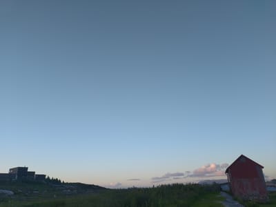 The image shows a rural landscape at dusk in Averøy, Norway. On the left, a dark, multi-segmented building complex is perched on a rocky, grassy incline, with a line of dark green trees behind it. On the right, a tall, red, corrugated metal barn stands at an angle. The sky is a pale blue with scattered, soft pink and white clouds on the horizon, suggesting the sun has recently set or is about to rise. The foreground consists of green grass and rocky terrain. The scene is devoid of people or any visible activity.
