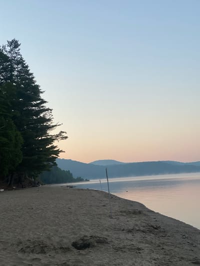 The image depicts a serene natural landscape, likely a beach on a body of water, during dawn or dusk. The foreground is a sandy beach with visible footprints and scattered debris, including branches and driftwood. To the left, a dense line of evergreen trees forms a natural barrier, their dark silhouettes contrasting with the lighter sky.
The middle ground features the calm water of a lake, with a thin layer of mist hovering just above the surface, particularly towards the distant shore. The water reflects the soft hues of the sky, transitioning from pale blue at the top to a gentle pink and peach near the horizon. In the distance, a chain of layered, hazy hills forms the backdrop, their outlines softened by the atmospheric conditions. Two thin, vertical poles, likely markers or part of a temporary structure, stand out on the beach and partially submerged in the water. No people are present in the scene.
The overall atmosphere is tranquil and untouched, suggesting a remote or natural setting. While the specific location is unknown, the environment is consistent with a lakeside scene in a forested region, potentially in the Canadian landscape, given the mention of Duhamel. However, without further information or context, a definitive link to Duhamel, Canada, cannot be made solely based on the visual elements of the image.