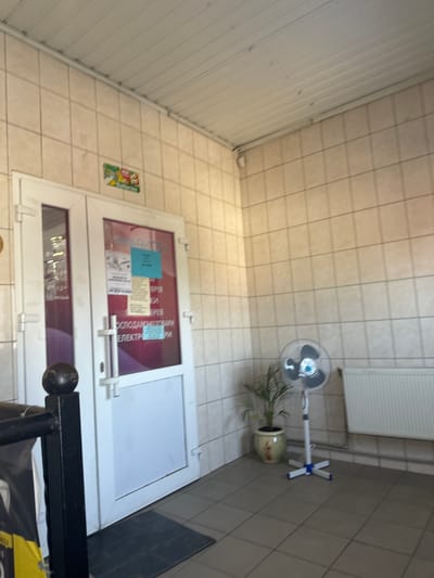 The image shows the exterior of a shop with a white door and a glass panel displaying various notices and signage. A black metal railing is partially visible in the foreground. To the right of the door, a floor fan stands next to a potted plant and a white radiator. The walls are tiled with beige square tiles. The ceiling appears to be a white ribbed panel. A small green and red sign with pictograms is affixed to the wall above the door. The location is indicated as being in Sarny, Ukraine. The overall impression is an outdoor entrance to a commercial establishment, possibly a small retail store. There are no people visible in the image. The lighting suggests daylight, possibly overcast.