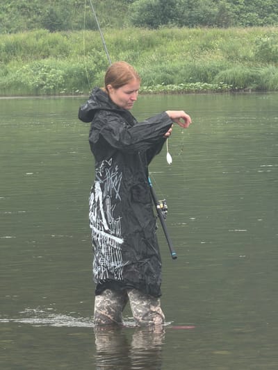 A person stands in the shallow water of a river, likely in Ust-Kabirza, Russia, facing right and holding a fishing rod. The person is wearing a black raincoat with white graffiti-style markings and camouflage waders. They are holding a fishing lure and line in their right hand, examining it. The river water is calm and green, with a lush, grassy bank visible in the background. The sky is overcast.