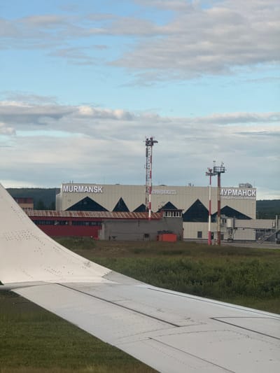 The image shows an aerial view of Murmansk Airport, located in Murmashi, Russia. The airport terminal building is visible with "MURMANSK" illuminated in large white letters. Below "MURMANSK", smaller text reads "ГОЛИЦА РУССКОЙ АРКТИКИ", which translates to "Gate to Russian Arctic". The terminal features a modern architectural design with triangular elements. Two tall red and white striped control towers are present, equipped with antennas and lights. A jet bridge extends from one side of the terminal. In the foreground, the white wing of an airplane is visible, occupying the lower left portion of the image. The ground around the airport is covered with green grass and various types of vegetation. In the background, a dense line of trees forms a forest. The sky is partly cloudy, with a mix of blue sky and white and gray clouds. There are no visible people clearly depicted in the image. The overall scene suggests an active airport environment.