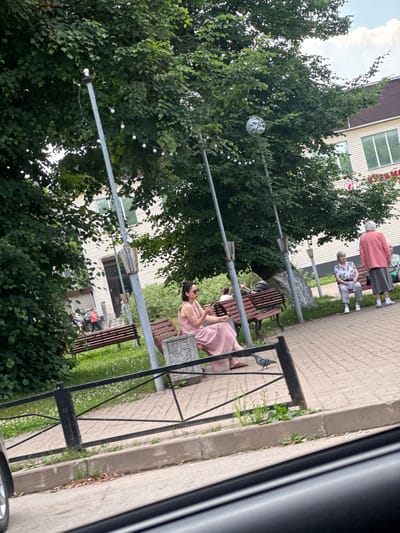 In Kuzmolovskiy, Russia, on a cloudy day, a woman in a pink dress is sitting on a bench, looking to her right. She is in the foreground of the image. In the background, other people are also seated on benches, some older individuals dressed in casual clothing. A building with light-colored siding and windows can be seen behind them. Trees with green foliage surround the area, and some string lights are visible in the trees. The scene appears to be a public park or outdoor seating area. The overall atmosphere seems relaxed and leisurely, with people enjoying the outdoors. There is no visible text in the image.